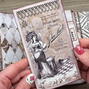 May include: A vintage-style journal with a decorative cover featuring a Victorian cabinet studio advertisement. The advertisement includes the text "Victoria Cabinet Studios" and "John Hart" with an illustration of a woman. The journal has a diamond pattern.