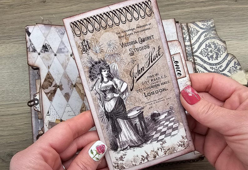 May include: A vintage-style journal with a decorative cover featuring a Victorian cabinet studio advertisement. The advertisement includes the text "Victoria Cabinet Studios" and "John Hart" with an illustration of a woman. The journal has a diamond pattern.