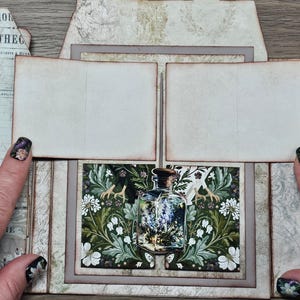 May include: A paper craft project featuring a central panel with a glass bottle illustration filled with lavender, surrounded by green foliage and white flowers. Above the bottle are two blank, rectangular, cream-colored paper flaps.