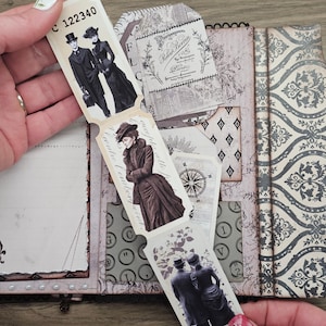 May include: A vintage-style journal with a decorative cover and pages. The journal features illustrations of people in Victorian-era clothing, a compass, and floral designs. The journal is held open, revealing the interior pages and attached tags.