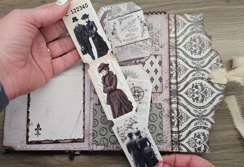 May include: A vintage-style journal with a decorative cover and pages. The journal features illustrations of people in Victorian-era clothing, a compass, and floral designs. The journal is held open, revealing the interior pages and attached tags.
