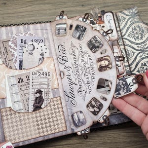 May include: A vintage-style scrapbook with a spiral binding, featuring pockets with old tickets and patterned paper. The cover has a striped design, and a decorative wheel with the text "Sawyer & Bird, Photographers" is visible.