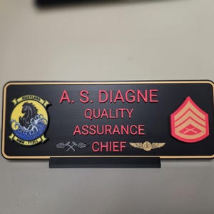 May include: A black desk nameplate with gold trim and a black stand. The nameplate features the name "A. S. DIAGNE" in red, along with the title "QUALITY ASSURANCE CHIEF". It includes military insignia.