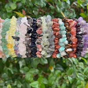 May include: A collection of gemstone chip bracelets in various colors, including pink, green, yellow, purple, black, and brown. The bracelets are strung together and displayed on a white cylindrical surface, with a backdrop of green foliage.