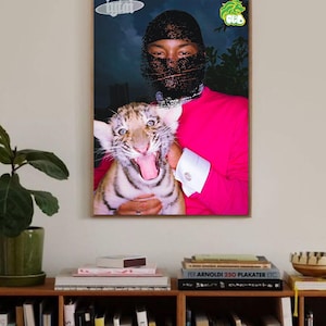 Puede incluir: Una impresión enmarcada que presenta a una persona con una camisa rosa y un pasamontañas negro sosteniendo un cachorro de tigre bostezando. La impresión tiene un fondo oscuro e incluye dos pegatinas en las esquinas superiores. La impresión se muestra encima de una estantería.