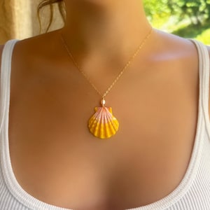 May include: A gold chain necklace featuring a scallop shell pendant with a gradient of yellow and pink hues. A small white pearl is positioned at the top of the shell where it connects to the delicate gold link chain, creating a simple accessory.