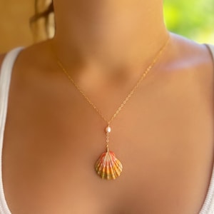 May include: A gold chain necklace featuring a small white pearl accent and a single seashell pendant. The seashell displays a gradient of orange, yellow, and pink hues with distinct vertical ridges, suspended from a delicate gold-toned metal chain.