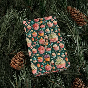 May include: A wrapped gift with a festive cupcake pattern on dark green wrapping paper. The design features cupcakes with various frosting colors, candles, and sprinkles. The gift is tied with twine and set against a backdrop of pine needles and pine cones.