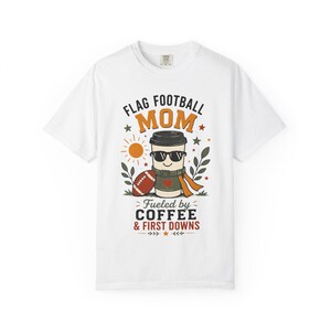 May include: A white short-sleeve t-shirt featuring a graphic of a coffee cup wearing sunglasses, next to a football. The text reads FLAG FOOTBALL MOM FUELED BY COFFEE & FIRST DOWNS, surrounded by small orange suns, stars, and green leaf illustrations.