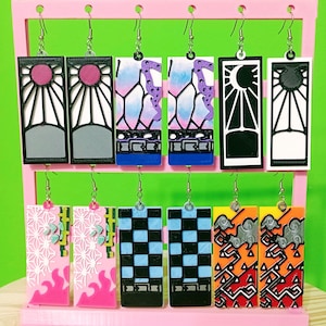 May include: A collection of rectangular earrings with various designs, including sunbursts, geometric patterns, and abstract shapes. The earrings feature a mix of colors like pink, blue, black, and orange, and are displayed on a pink earring stand.