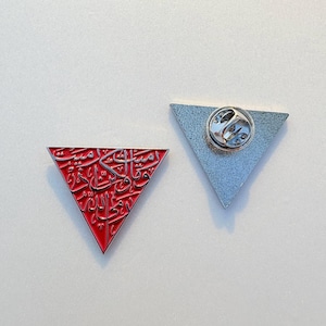 May include: Two triangular enamel pins. One is red with silver Arabic script, the other is silver with a pin back. The red pin has a raised silver border. The pins are likely for clothing or accessories.
