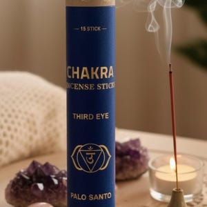 Op de afbeelding: Een blauwe tube met Chakra wierookstokjes met gouden letters en een symbool van het derde oog. Een brandende wierookstok geeft rook af, naast een brandende kaars en kristallen. De tube leest "Palo Santo" en "Handmade in Nepal."