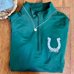 Puede incluir: Una camiseta deportiva verde oscuro con cremallera de un cuarto, una herradura blanca y la palabra "Colts" impresa en la parte delantera. Un collar de oro con un pequeño colgante está encima de la camisa. Una pequeña planta en maceta está en una cesta tejida.