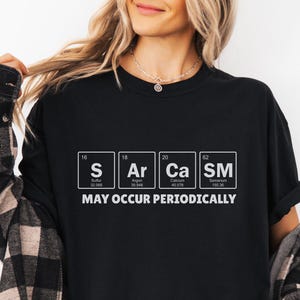 May include: Black long-sleeve t-shirt with the periodic table elements S, Ar, Ca, and Sm, spelling out "Sarcasm". The text "May Occur Periodically" is below the elements. The shirt is a casual style.