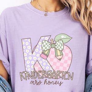 May include: A light purple t-shirt featuring a graphic design for kindergarten. The design includes the letters "K" and "G" with floral and checkered patterns, an apple, and the words "Kindergarten" and "mrs honey" in a playful font.