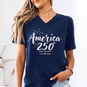 May include: A navy blue V-neck t-shirt featuring white screen-printed text that reads America 250th Anniversary 1776 2026, accompanied by a small star graphic design above the word America and a single star between the years 1776 and 2026.