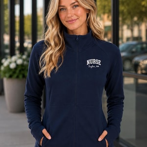 May include: A navy blue zip-up jacket with a high collar and long sleeves featuring thumb holes. The left chest area displays white embroidered text that reads NURSE followed by the name Taylor, RN in a smaller script font below the word.