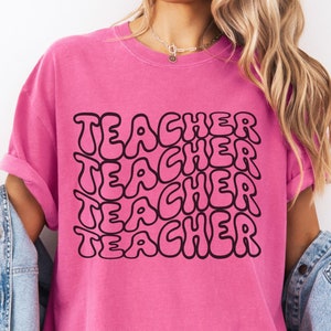 May include: Pink t-shirt with the word "TEACHER" repeated in a retro, bubble-style font.