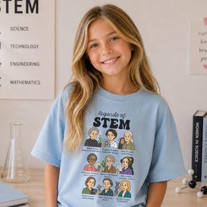 May include: A light blue t-shirt featuring a graphic print titled "legends of STEM" with illustrations of nine historical female scientists, including Marie Curie and Jane Goodall, each labeled with their name and field of study in black text.
