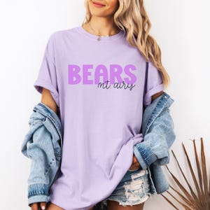 K&ouml;nnte beinhalten: Ein hellviolettes Kurzarm-T-Shirt mit dem Wort BEARS in gro&szlig;en, fetten, violetten Gro&szlig;buchstaben und dem Text mt. airy darunter in einer kleineren, schwarzen Schreibschrift. Das T-Shirt hat einen Rundhalsausschnitt und eine lockere Passform.
