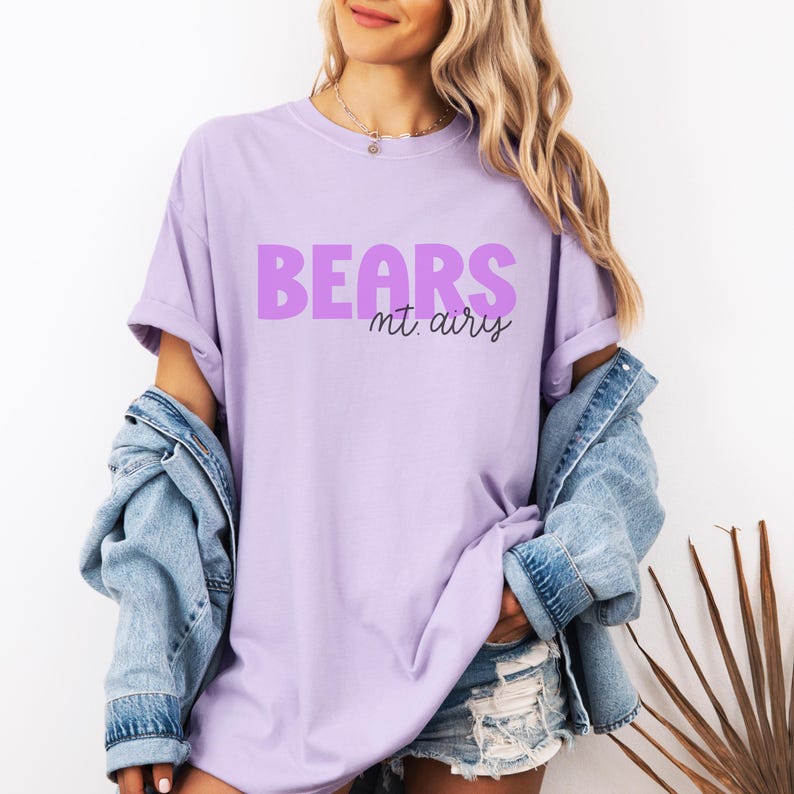 K&ouml;nnte beinhalten: Ein hellviolettes Kurzarm-T-Shirt mit dem Wort BEARS in gro&szlig;en, fetten, violetten Gro&szlig;buchstaben und dem Text mt. airy darunter in einer kleineren, schwarzen Schreibschrift. Das T-Shirt hat einen Rundhalsausschnitt und eine lockere Passform.