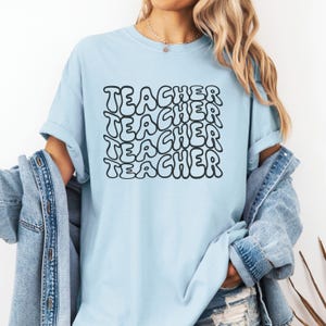 May include: A light blue t-shirt with the word "TEACHER" repeated in a black, bubble-like font.