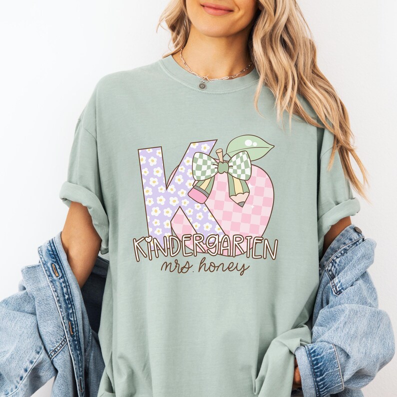 May include: A light green t-shirt with a graphic that says "Kindergarten mrs. honey." The "K" is lavender with white flowers, and the "6" is pink and white checkered. A green apple with a bow and pencil is in the middle.