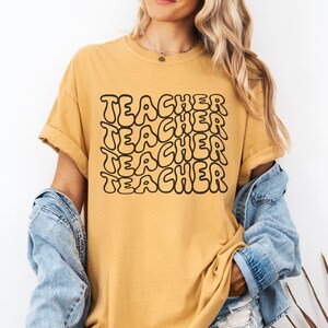 May include: A mustard yellow t-shirt with the word "TEACHER" repeated in a black, bubble-like font.