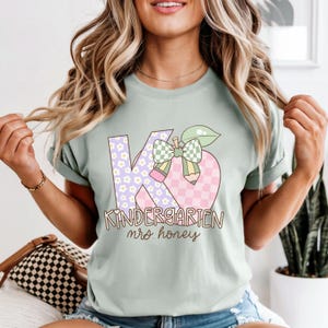 May include: A light green t-shirt featuring a colorful graphic design. The design includes the letters "K" and "6" with floral and checkered patterns, a bow, and a pencil. The words "Kindergarten mrs. honey" are printed below the graphic.