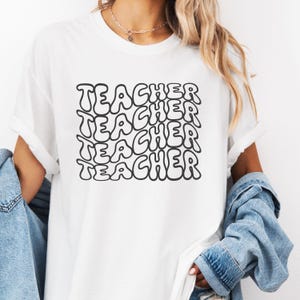May include: White t-shirt with the word "TEACHER" repeated four times in a black, bubble-like font.