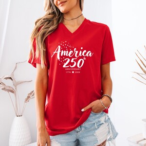 May include: A red V-neck t-shirt featuring white graphic text that reads America 250th Anniversary 1776 2026 with a star design above the word America. The shirt has short sleeves and a standard fit, showcasing a celebratory design for the nation.