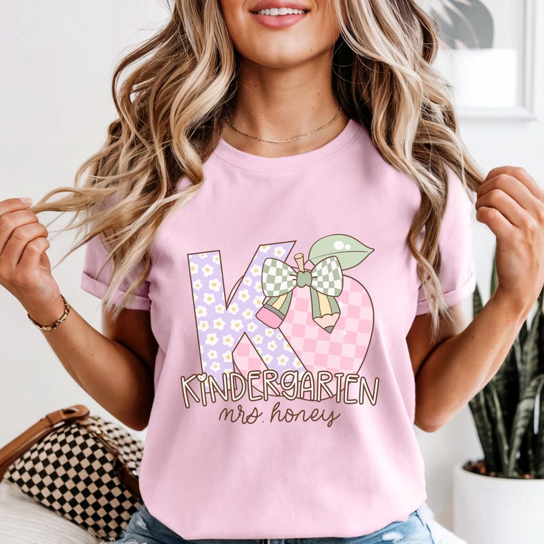 May include: A light pink t-shirt featuring a graphic design for kindergarten. The design includes the letters "K" and "6" with a floral pattern, a checkered apple, a bow, and the words "Kindergarten mrs. honey".
