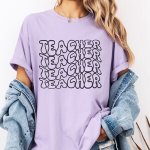May include: A lavender t-shirt with the word "TEACHER" repeated in a retro, bubble-like font.