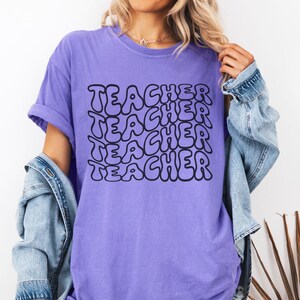 May include: Purple t-shirt with the word "TEACHER" repeated in a retro, bubble-letter style.