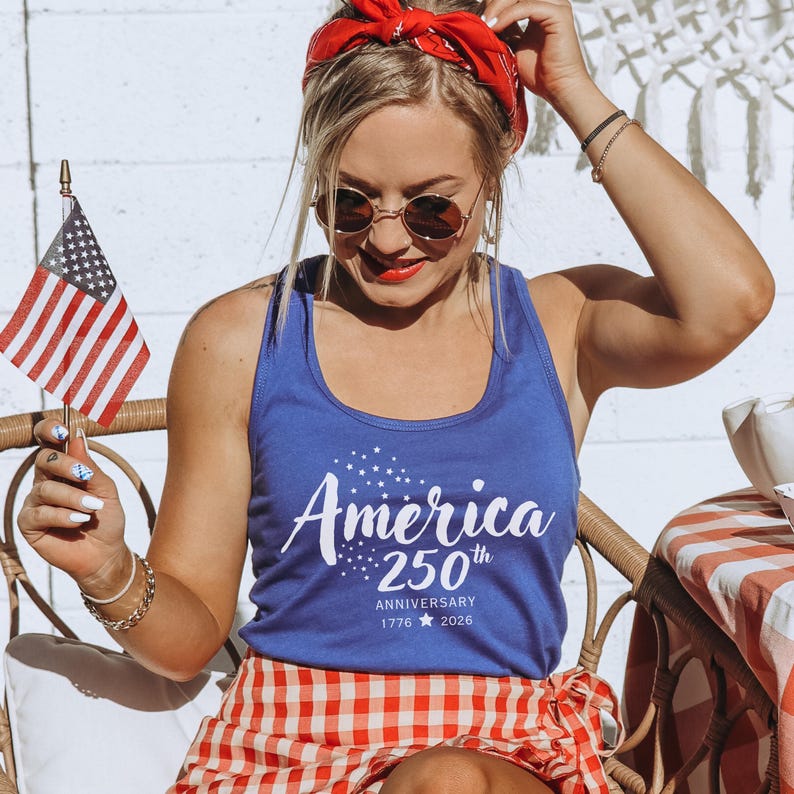 Pu&ograve; includere: Una canotta blu con la scritta "America" e "250th Anniversary 1776-2026" stampata in bianco. La persona tiene in mano una piccola bandiera americana e indossa una bandana rossa e una gonna a scacchi rossi e bianchi.