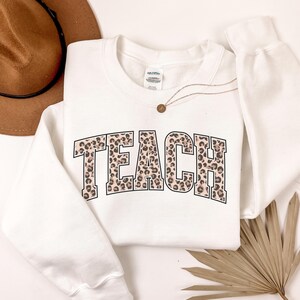 Op de afbeelding: Een witte sweatshirt met het woord "TEACH" in grote letters met luipaardprint. Een gouden ketting en een bruine hoed zijn zichtbaar op de achtergrond. De sweatshirt heeft een ronde hals.