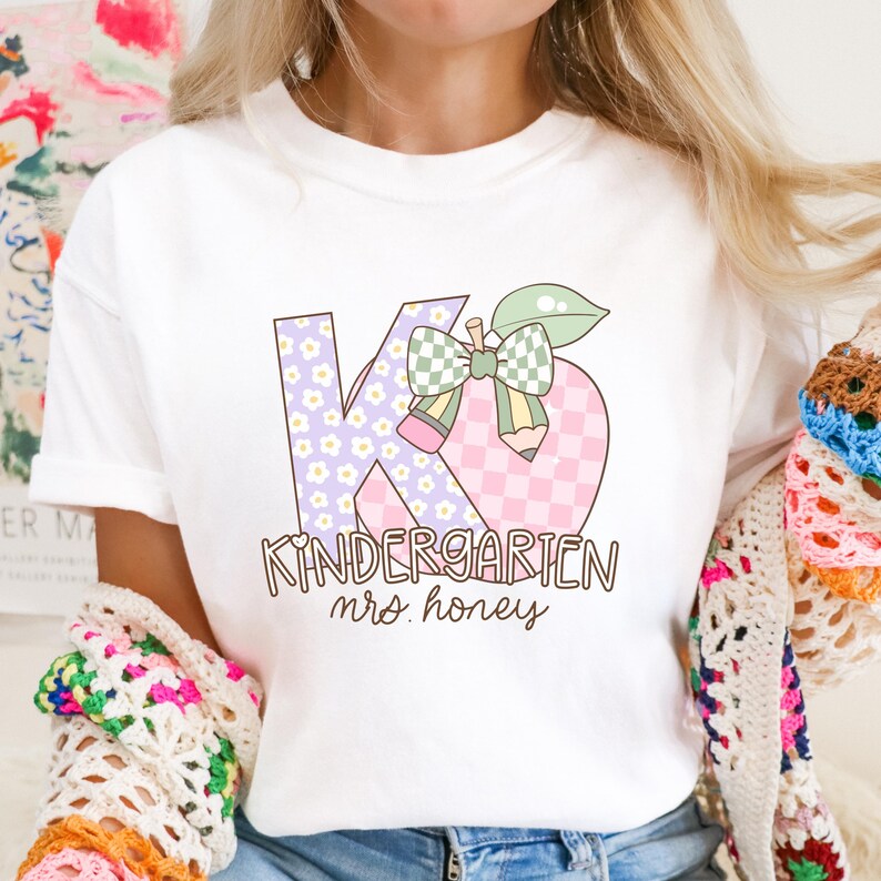 May include: White t-shirt with a colorful graphic design. The design features the letters "KG" in a floral pattern and a pink and white checkered pattern, along with the word "Kindergarten" and "mrs. honey". A pencil and bow complete the design.