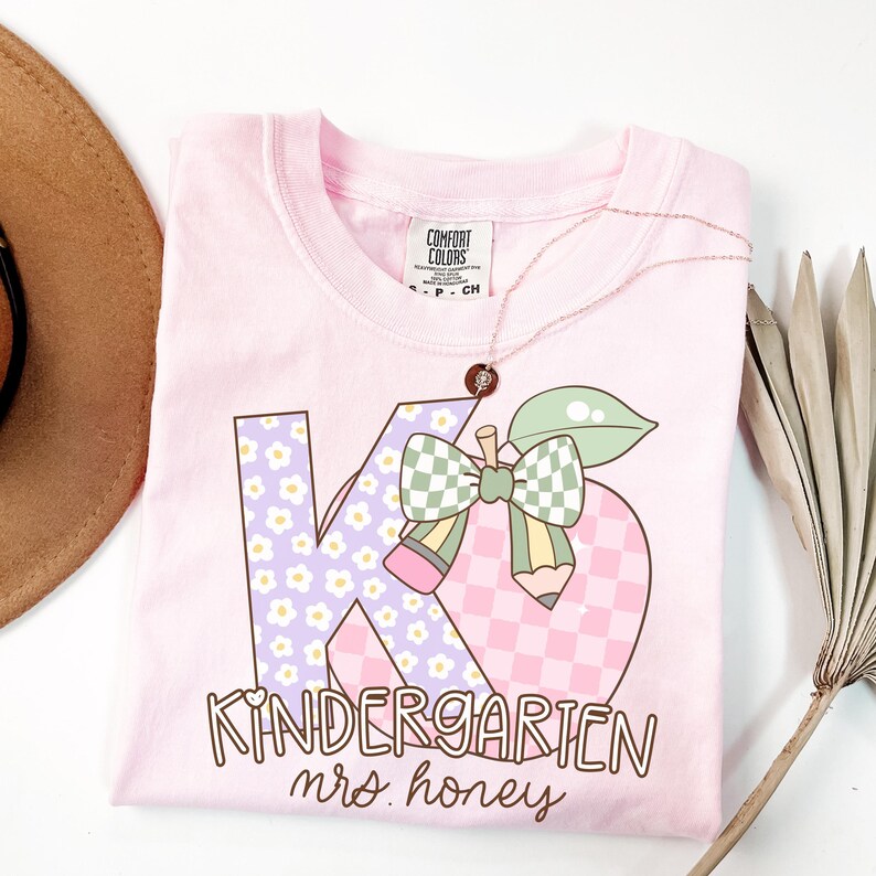 May include: A light pink Comfort Colors t-shirt with a graphic design for kindergarten. The design includes the letters "K" and "6" with floral and checkered patterns, a pencil, and the words "Kindergarten mrs. honey". A rose gold necklace is draped over the shirt.