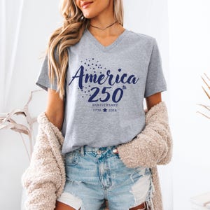May include: A heather grey V-neck t-shirt featuring dark blue text that reads America 250th Anniversary 1776-2026 with a small star graphic. The design includes a cluster of small stars above the word America and a single star between the years.