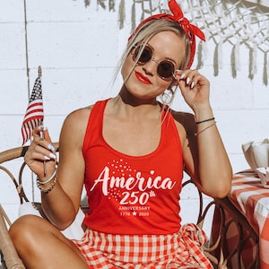 Pu&ograve; includere: Canotta rossa con la scritta "America" e "250th Anniversary 1776-2026" stampata in bianco. La persona indossa una bandana rossa e occhiali da sole rotondi. Una piccola bandiera americana &egrave; tenuta in mano.