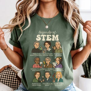 May include: Green t-shirt featuring the text "legends of STEM" and illustrations of notable women in science. The shirt includes names like Marie Curie, Rosalind Franklin, and Jane Goodall, with their respective fields of study listed below each portrait.