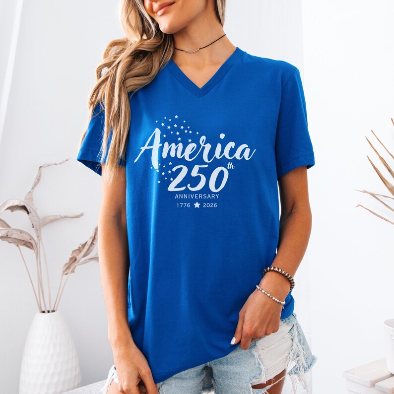 May include: A royal blue V-neck t-shirt featuring white screen-printed text that reads America 250th Anniversary 1776 2026 with a small star icon between the years and a cluster of small stars above the word America on the front of the shirt.