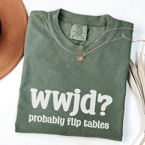 May include: An olive green t-shirt with the text "wwjd? probably flip tables" in white. The shirt has a crew neck and short sleeves. A gold necklace with a pendant is draped across the shirt.