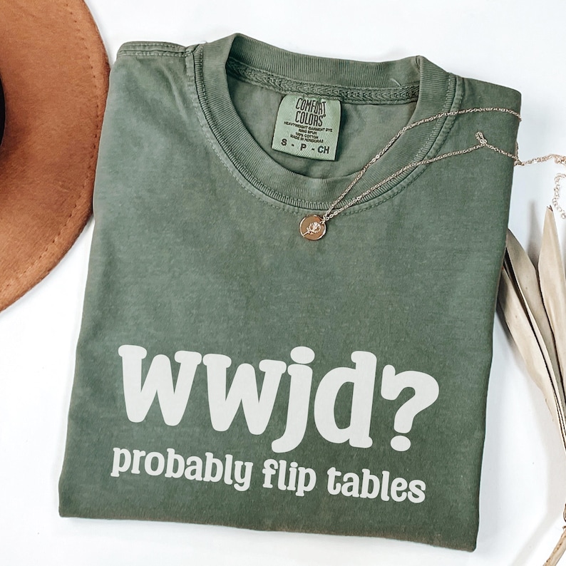 May include: An olive green t-shirt with the text "wwjd? probably flip tables" in white. The shirt has a crew neck and short sleeves. A gold necklace with a pendant is draped across the shirt.