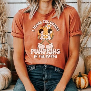 May include: A woman wearing a burnt orange t-shirt with a graphic of a smiling pumpkin with the text "I teach the cutest pumpkins in the patch".