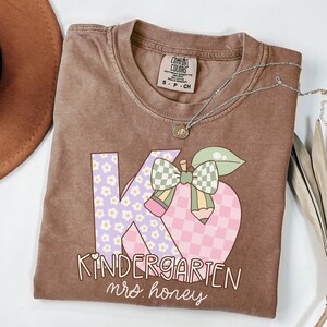 May include: A brown Comfort Colors t-shirt with a graphic design for a kindergarten teacher. The design features the letters "K" and "G" with floral and checkered patterns, a pencil, and the words "Kindergarten mrs. honey."