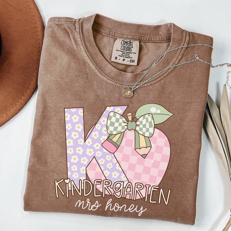 May include: A brown Comfort Colors t-shirt with a graphic design for a kindergarten teacher. The design features the letters "K" and "G" with floral and checkered patterns, a pencil, and the words "Kindergarten mrs. honey."