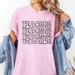 May include: A pink t-shirt with the word "TEACHER" repeated four times in black, outlined letters. The letters are in a bubble-like font.