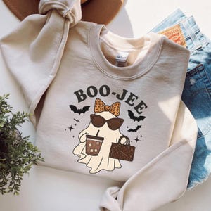May include: Beige crewneck sweatshirt with the text "BOO-JEE" above a cartoon ghost wearing sunglasses and a leopard print bow. The ghost is holding a coffee cup and a purse. Black bats and stars surround the ghost.