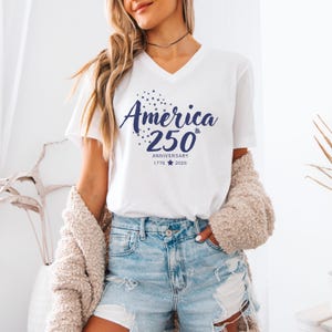 May include: A white short-sleeved V-neck t-shirt featuring dark blue text that reads America 250th Anniversary 1776-2026, accompanied by a cluster of small stars and a single star icon between the years, printed on the front of the garment.